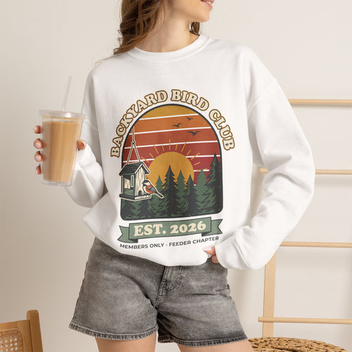 Crewneck sweatshirt with retro 'Backyard Bird Club Est. 2026' badge design featuring a sunset, pine trees and a chickadee at a bird feeder